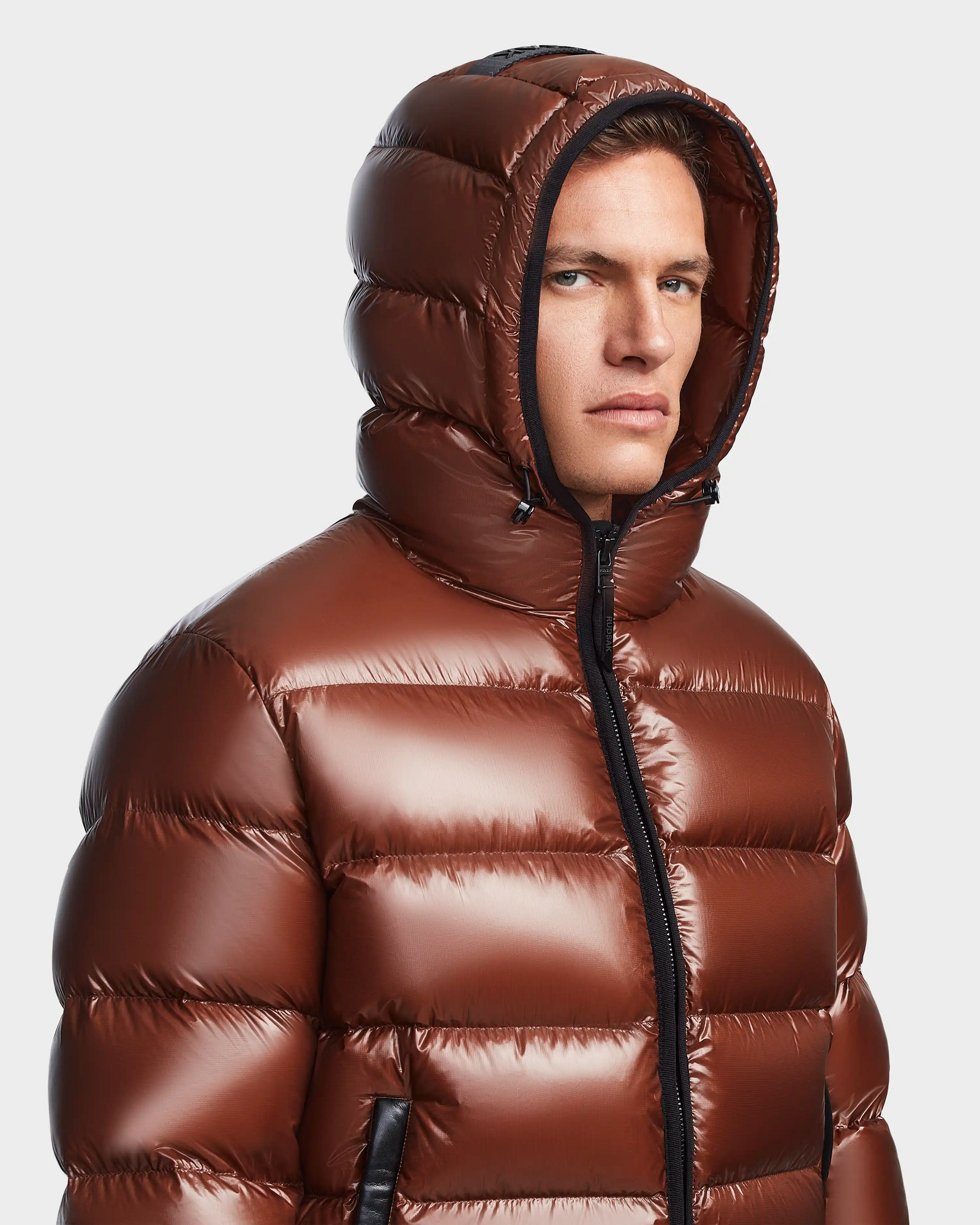 A man wears the Rudsak DEMIAN S - M123508 Cohiba, a shiny brown hooded mens down puffer, looking to the side with a neutral expression against a plain background. The water- and wind-resistant jacket blends style and function.