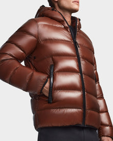 A person wears the Rudsak DEMIAN S - M123508 Cohiba, a shiny brown hooded down puffer jacket with black zipper and side pockets. Warm, quilted, water- and wind-resistant, its shown from mid-chest up as the wearer looks slightly to the side.