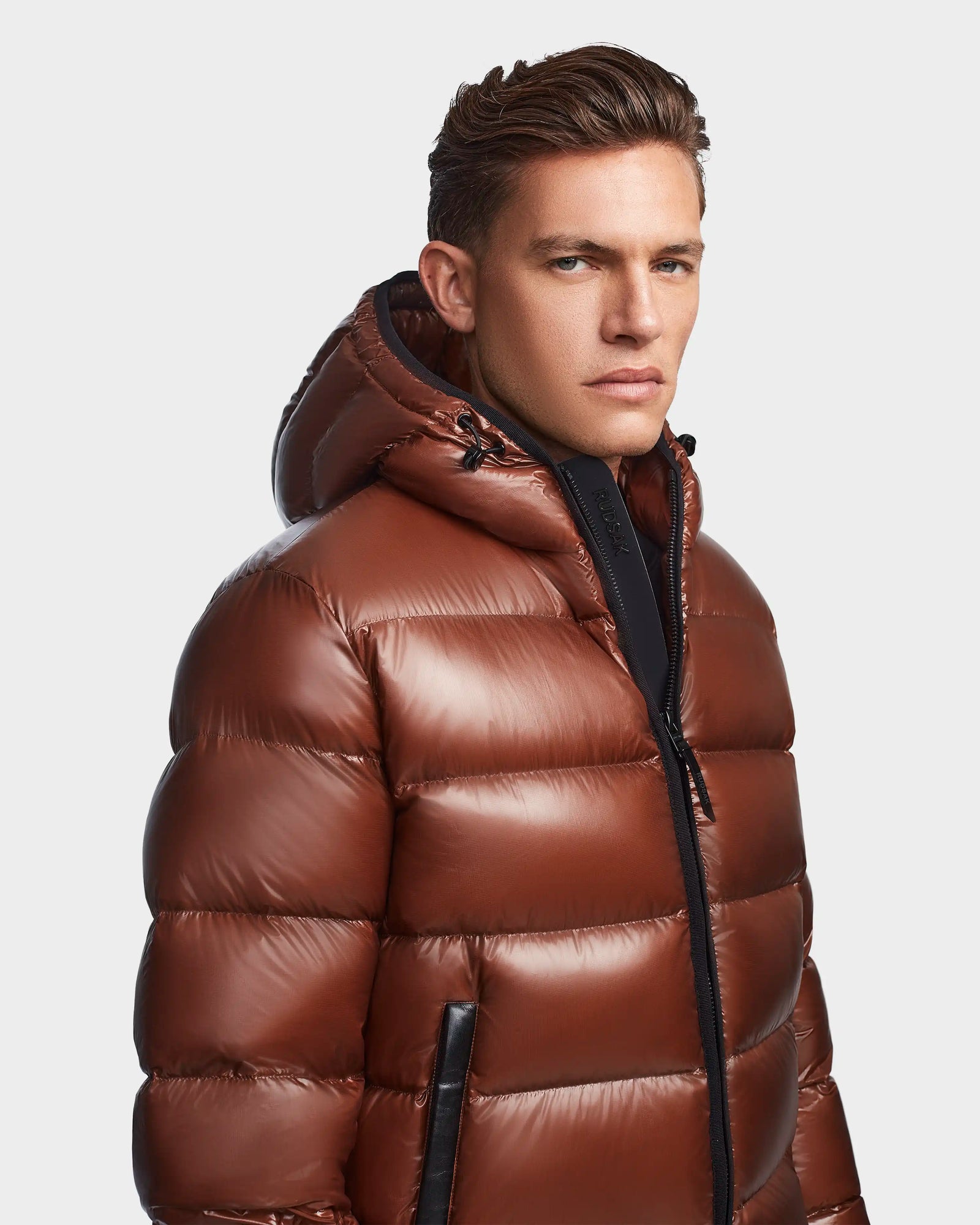 A man wearing the Rudsak DEMIAN S - M123508 Cohiba shiny brown, water and wind-resistant hooded down puffer faces slightly to the side, looking at the camera with a neutral expression against a plain background.
