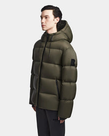 A man wearing the Rudsak DANIEL M - M123510 Light Olive mens hooded down puffer with a black zipper and upper arm patch stands facing left against a plain light background.
