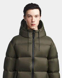 A person with short dark hair models the Rudsak DANIEL M - M123510 Light Olive men’s hooded down puffer coat, standing facing forward against a plain light background.