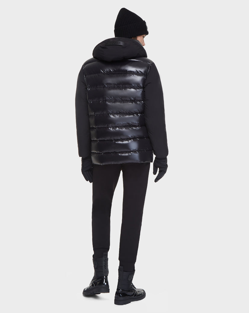 A person faces away, wearing the Rudsak DUSK - M121506 BLACK men's shiny black puffer jacket with waterproof zippers, along with a black beanie, gloves, pants, and shoes against a plain white background.