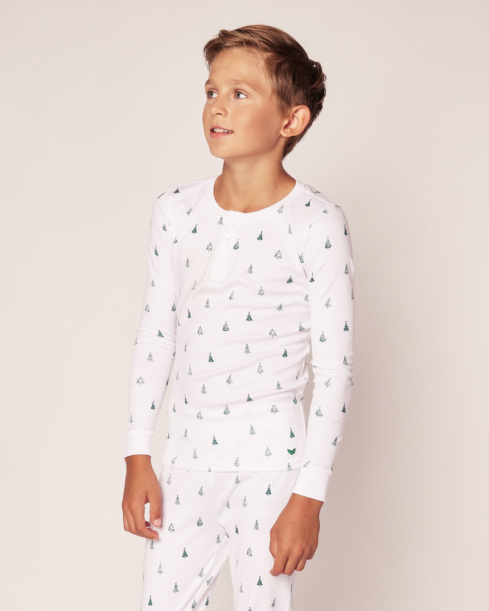 A child in Petite Plumes Kids Pima Snug Fit Pajama Set in Evergreen Trees, made of soft Pima cotton, stands against a plain background looking to the side. The long sleeve top and pant set exudes a light and neutral tone.