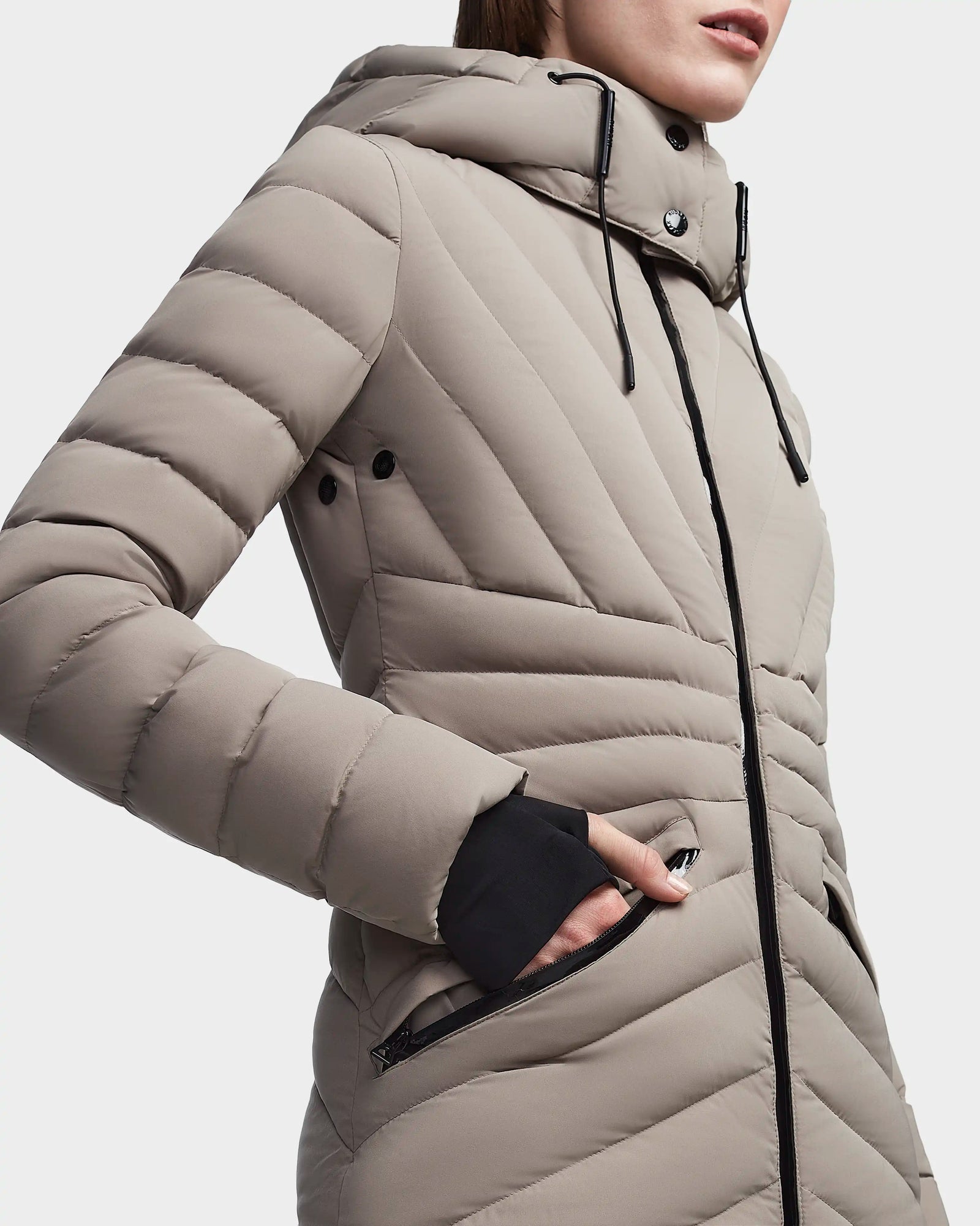 A person wears the Rudsak HANNA - 8123562 Cement, a light brown, RDS-certified quilted down jacket with black zippers, one hand in the pocket, shown in a close-up highlighting the upper body and jacket details.