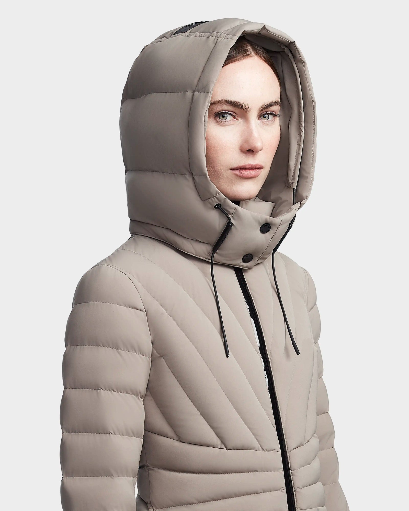 A woman models the Rudsak HANNA - 8123562 Cement, a light taupe hooded down jacket with black drawstrings. Featuring horizontal and diagonal quilting, this winter coat offers stylish warmth against a plain light background.