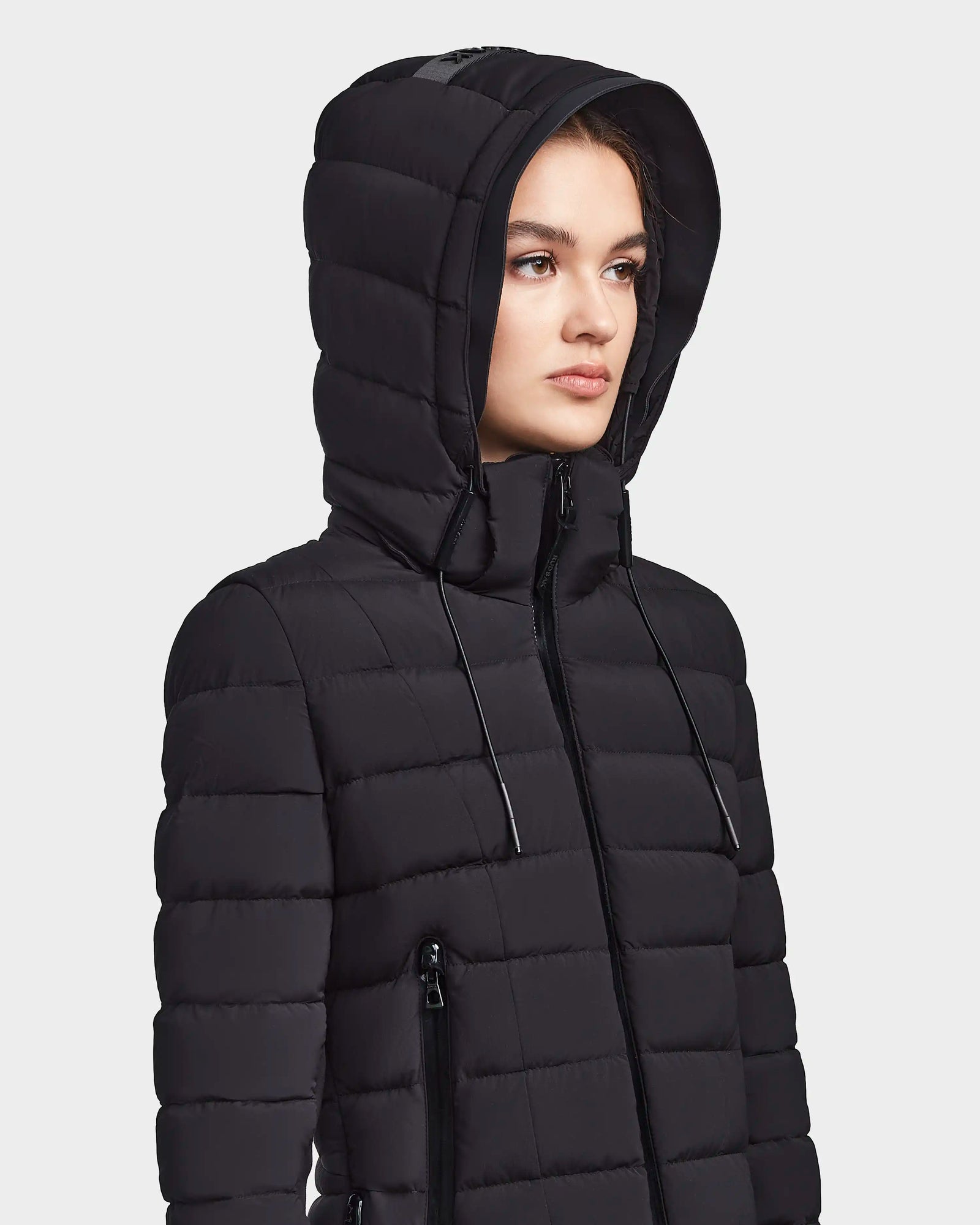 A woman wears the RUDSAK MELISSA - 8122551 BLACK weatherproof down jacket with a hood, zippered pockets, and high neck, looking forward with a neutral expression against a plain background.