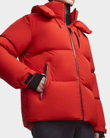 A person wears the Rudsak VINCENT M - 6123510 Orange men's down puffer jacket, featuring ultra-warm insulation, zippered pockets, and a water-repellent finish. The jacket is partially unzipped, showing a black inner layer; only the torso and lower face are visible.