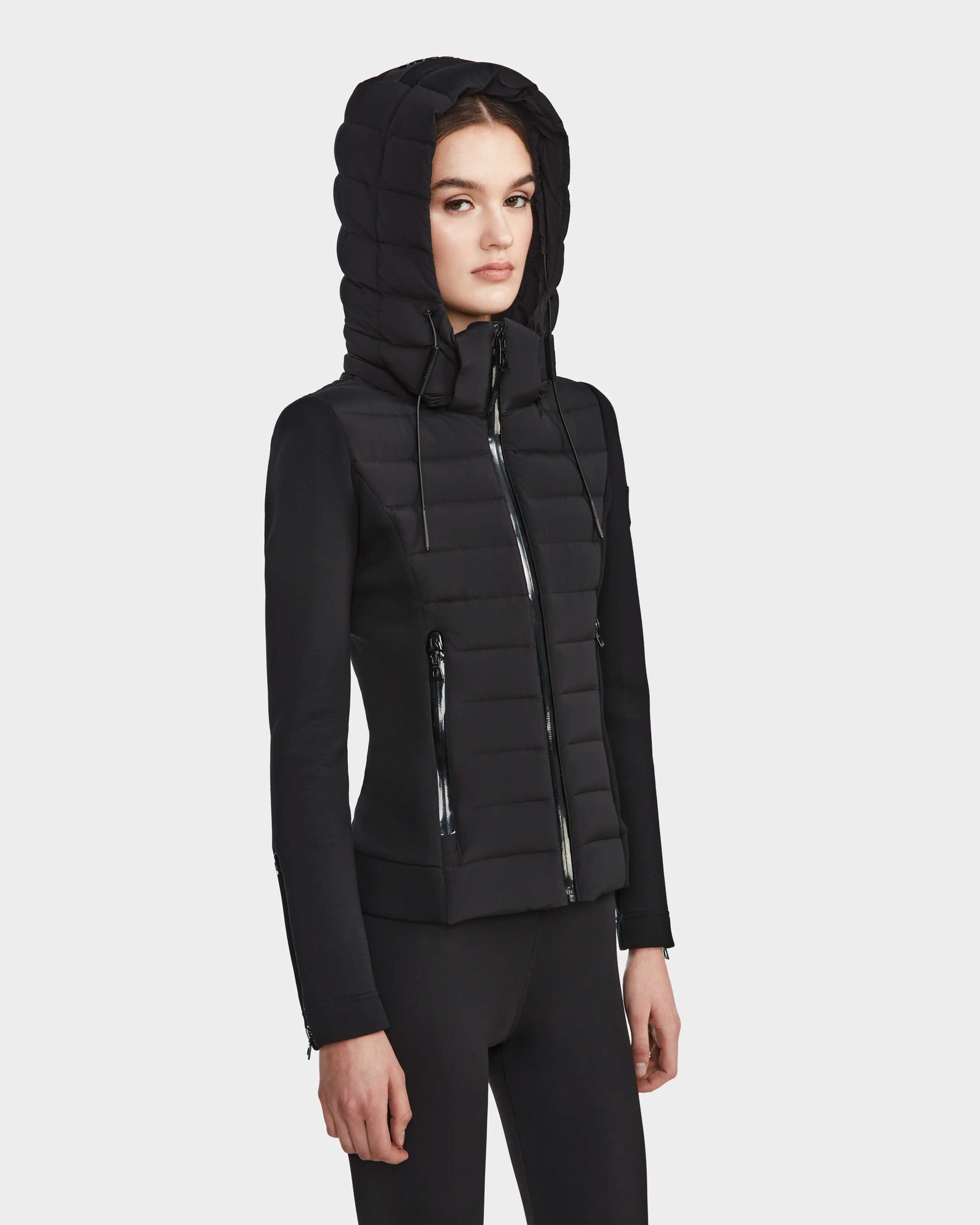 A woman in the RUDSAK MABS - 8122553 BLACK fitted hooded puffer jacket with zippered pockets and black leggings stands against a plain white background.
