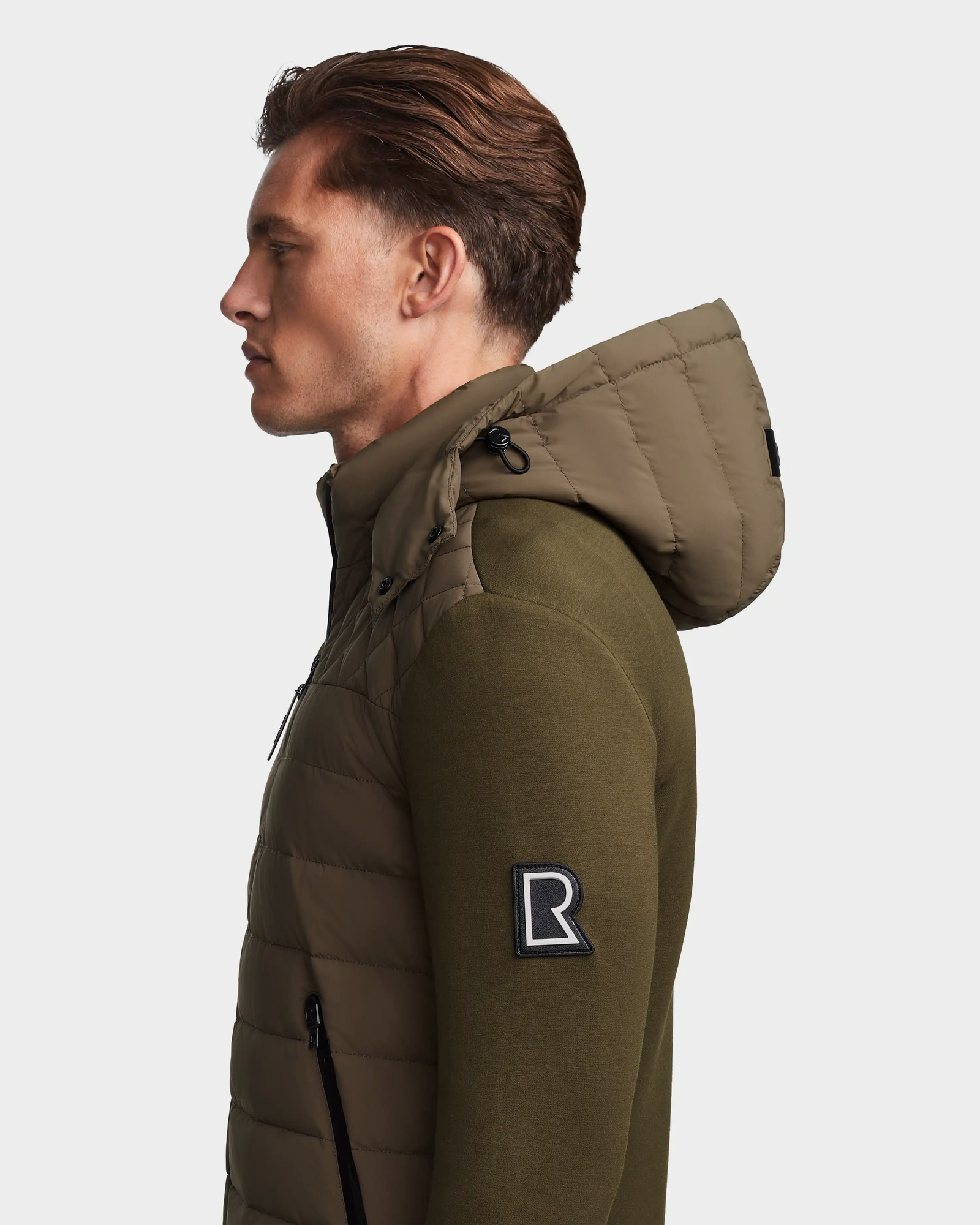 A man with short, styled brown hair wears the Rudsak LAURENZO - M123514 Light Olive jacket with a hood and black R patch on the sleeve, shown in profile against a plain white background.