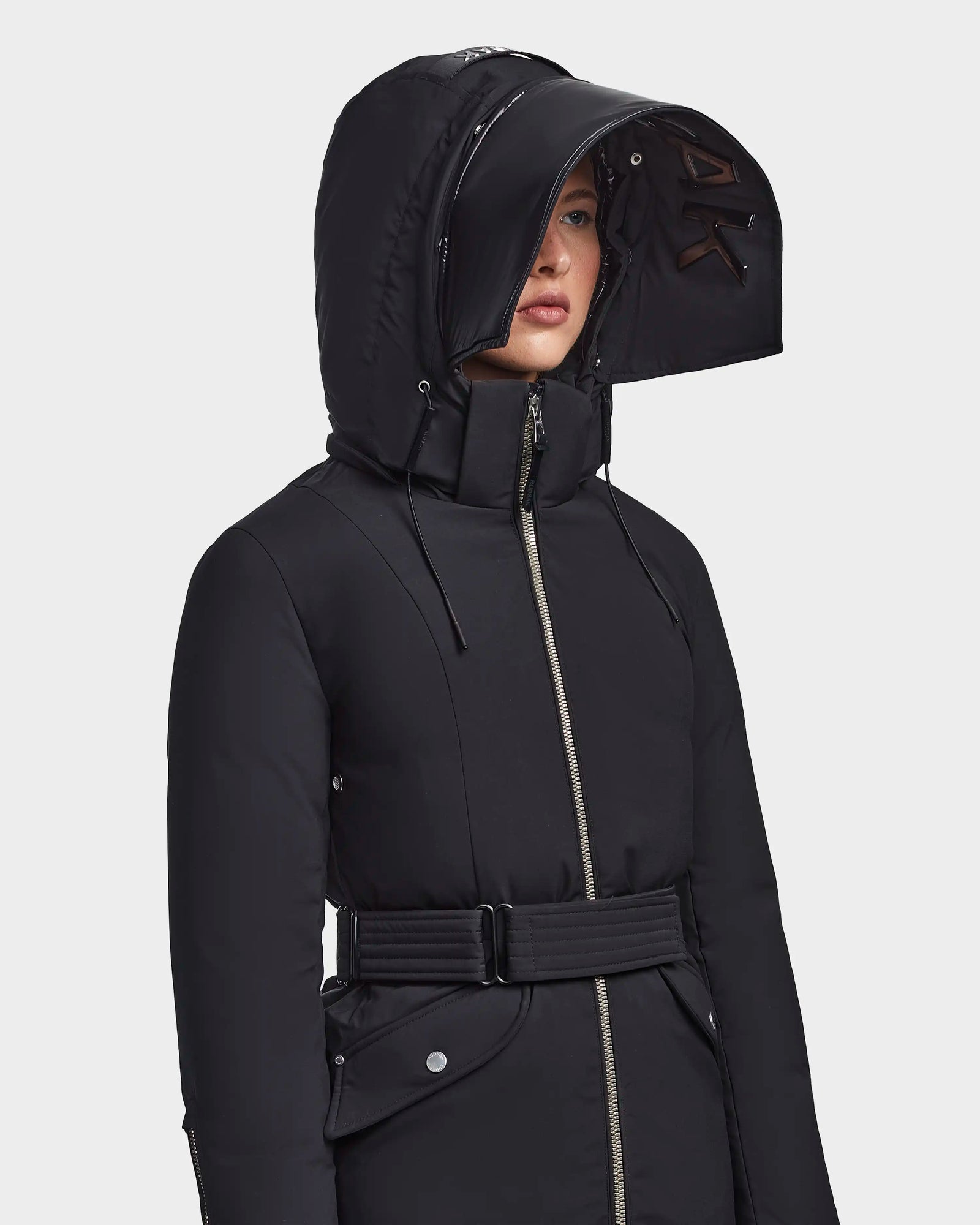 A woman stands against a light background wearing the RUDSAK MILENA - 8122536 BLACK, a long recycled Stormshell winter coat with a structured hood, front zipper, and belted waist.