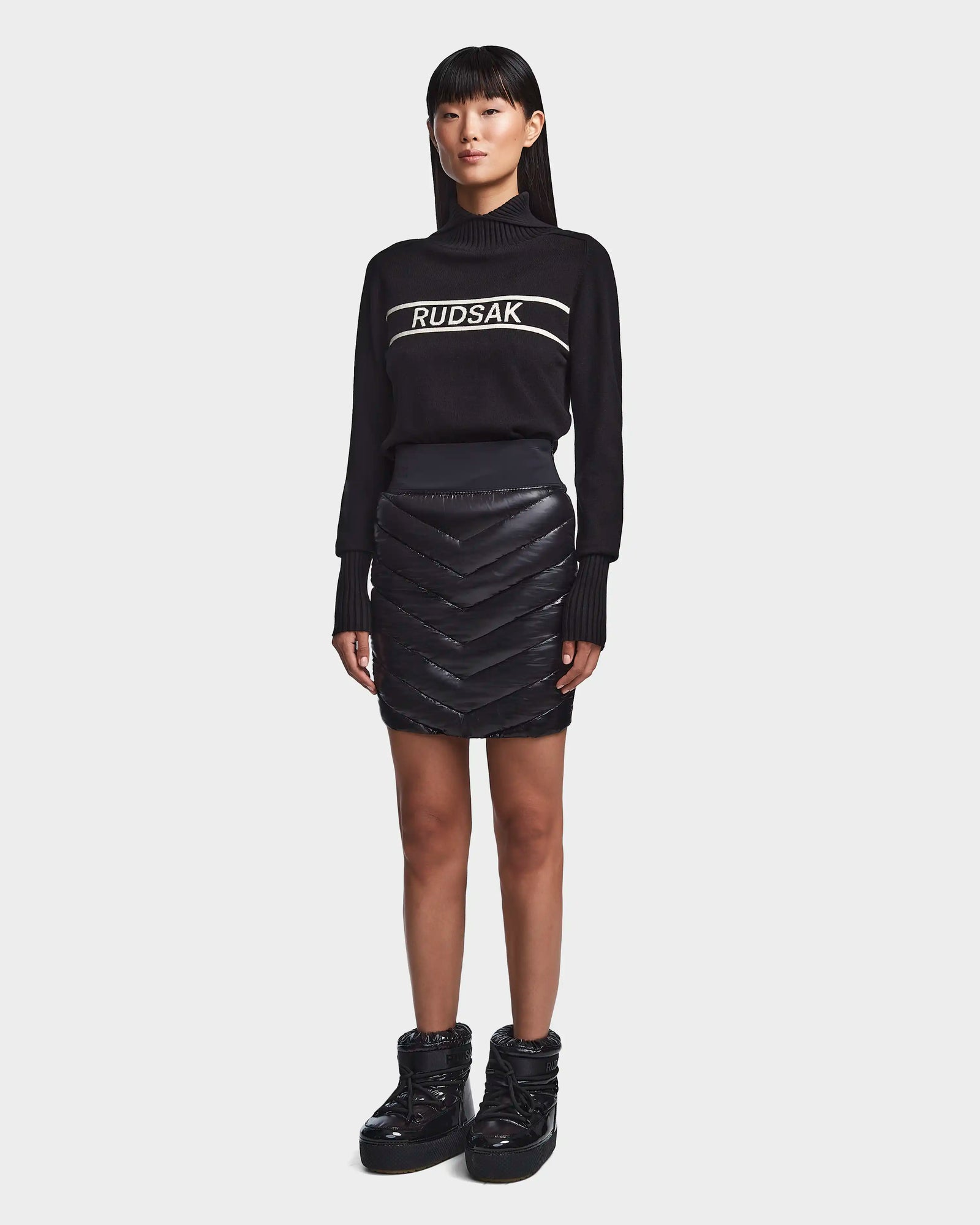 A woman faces forward in a black Rudsak turtleneck sweater, the MARZI - 8523528 Black chevron quilted winter skirt, and black quilted ankle boots, posed against a plain light gray background.