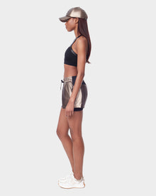 A woman stands in profile wearing the RYLEE - 8523001 METALLIC CHOCOLAT running shorts with spandex inner short by Rudsak, a metallic cap, black sports bra, and white sneakers against a plain white background.
