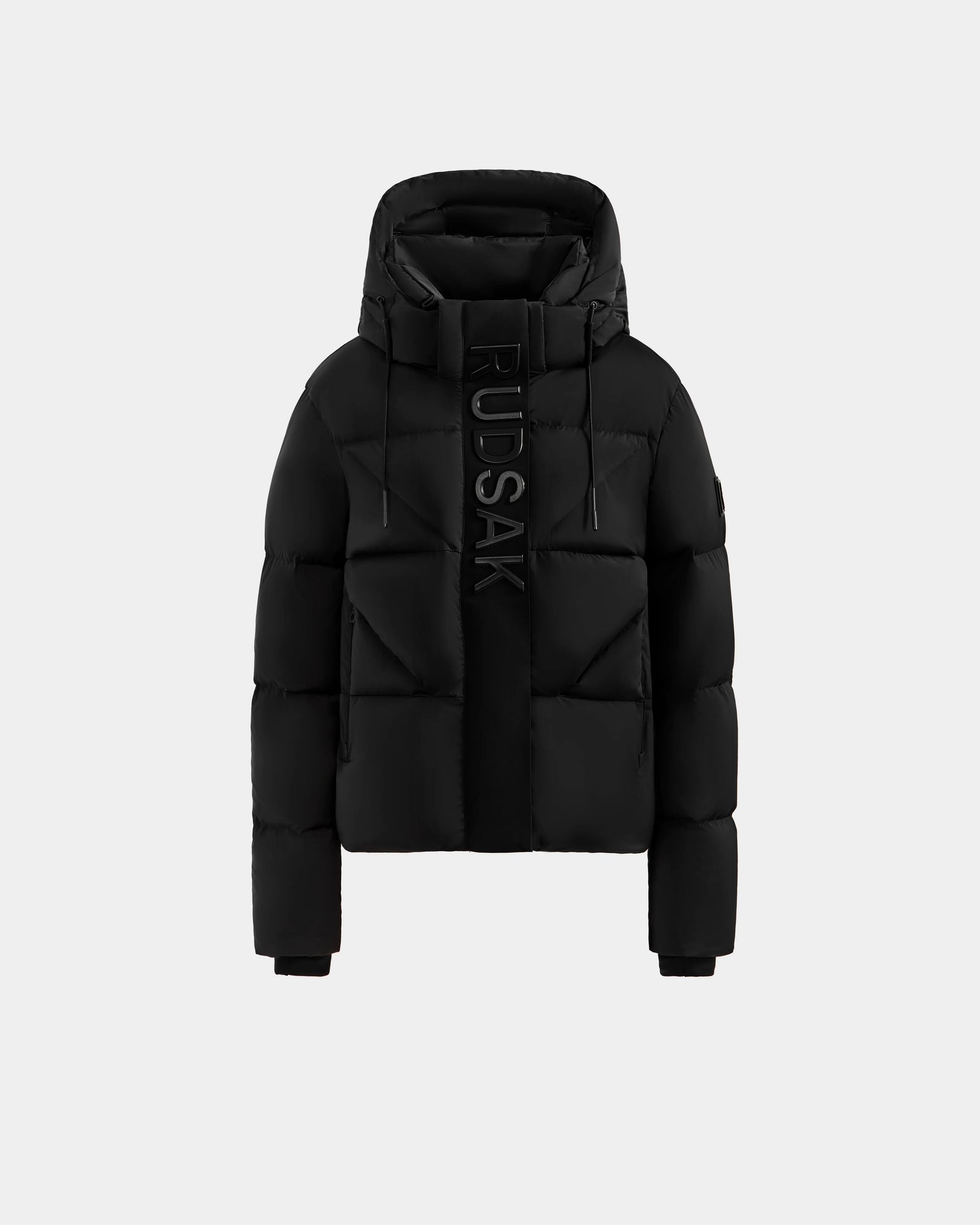 The Rudsak STEFANIE M - L125526 Black is a black puffer jacket with a hood, front zipper, and large vertical “RUDSAK” branding, shown against a light gray background.