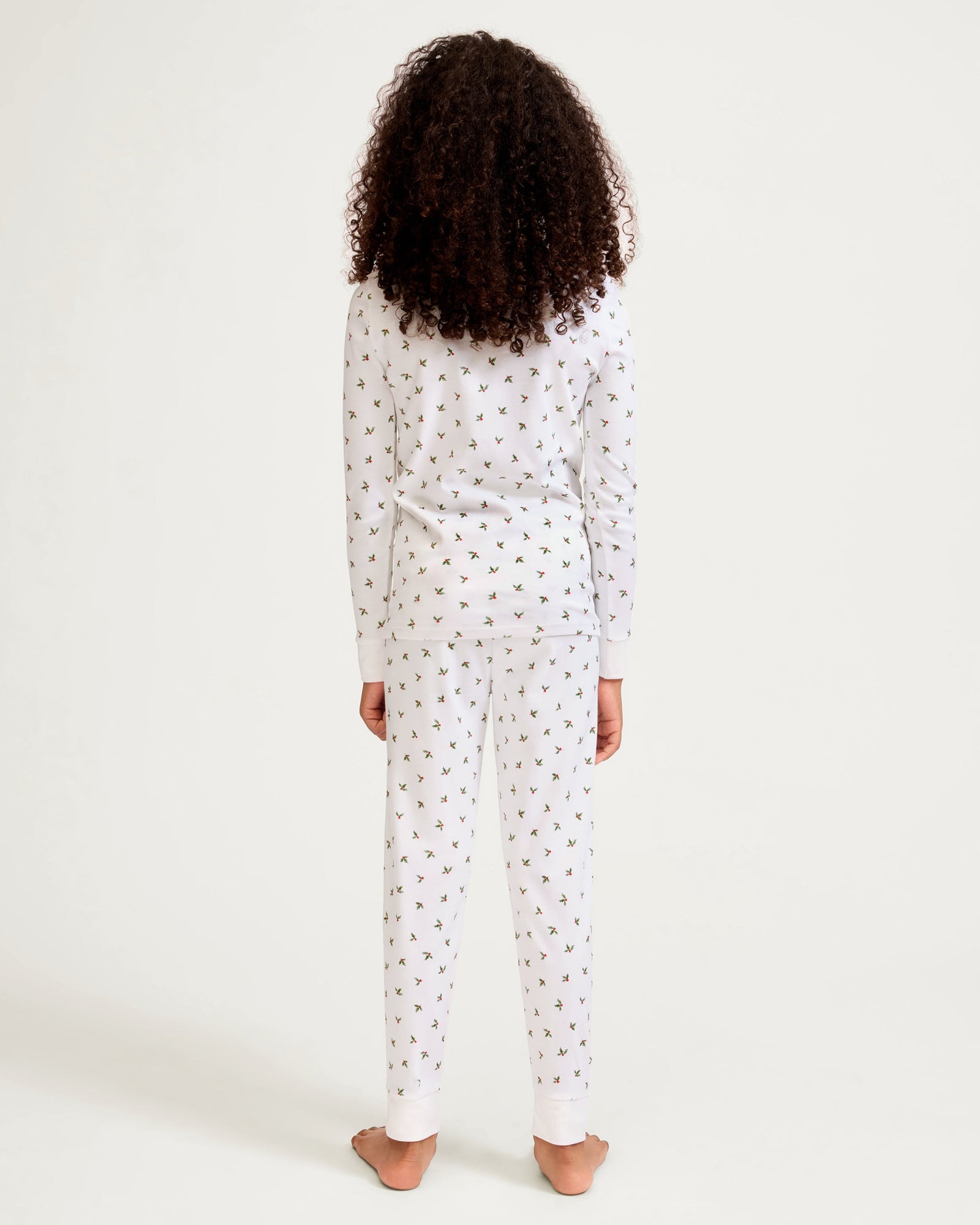 Kid's Pima Snug Fit Pajama Set in Mistletoe Moments: a two-piece white pajama set for children with a long-sleeve top, pants, green and red holly pattern, red scalloped neckline, and a red bow at the collar.