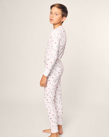 A young boy in a Petite Plume Kids Pima Snug Fit Pajama Set, Apres Ski design, with long sleeves and colorful illustrations, stands barefoot on a plain backdrop, glancing over his shoulder at the camera.