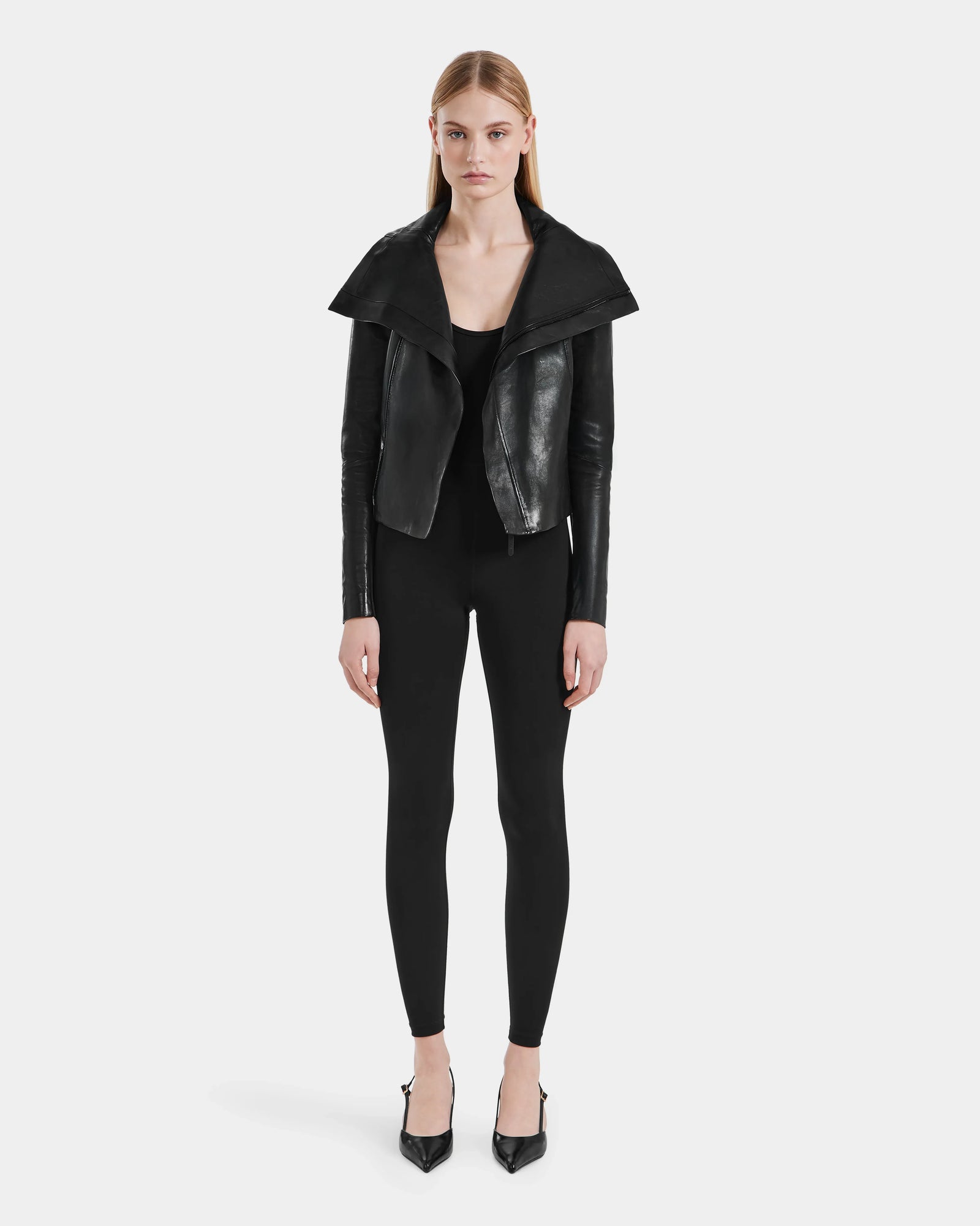 A woman with straight light brown hair and a neutral expression faces forward, wearing the AVRIL - 8124561 Black leather jacket by Rudsak over a black top and fitted pants, paired with black pointed-toe heels.
