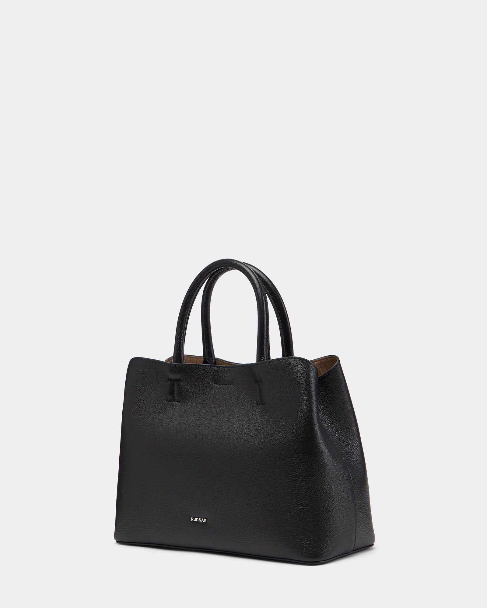The Rudsak CATIRA S - 8325006 Black is a women’s leather tote bag featuring two handles, minimal stitching, a subtle gold logo near the bottom, and a removable shoulder strap, displayed on a plain white background.