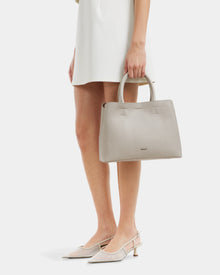 A woman in a white dress and pointed-toe heels holds the Rudsak CATIRA S - 8325006 Vanilla pebble leather tote bag with top handles, standing against a plain white background.