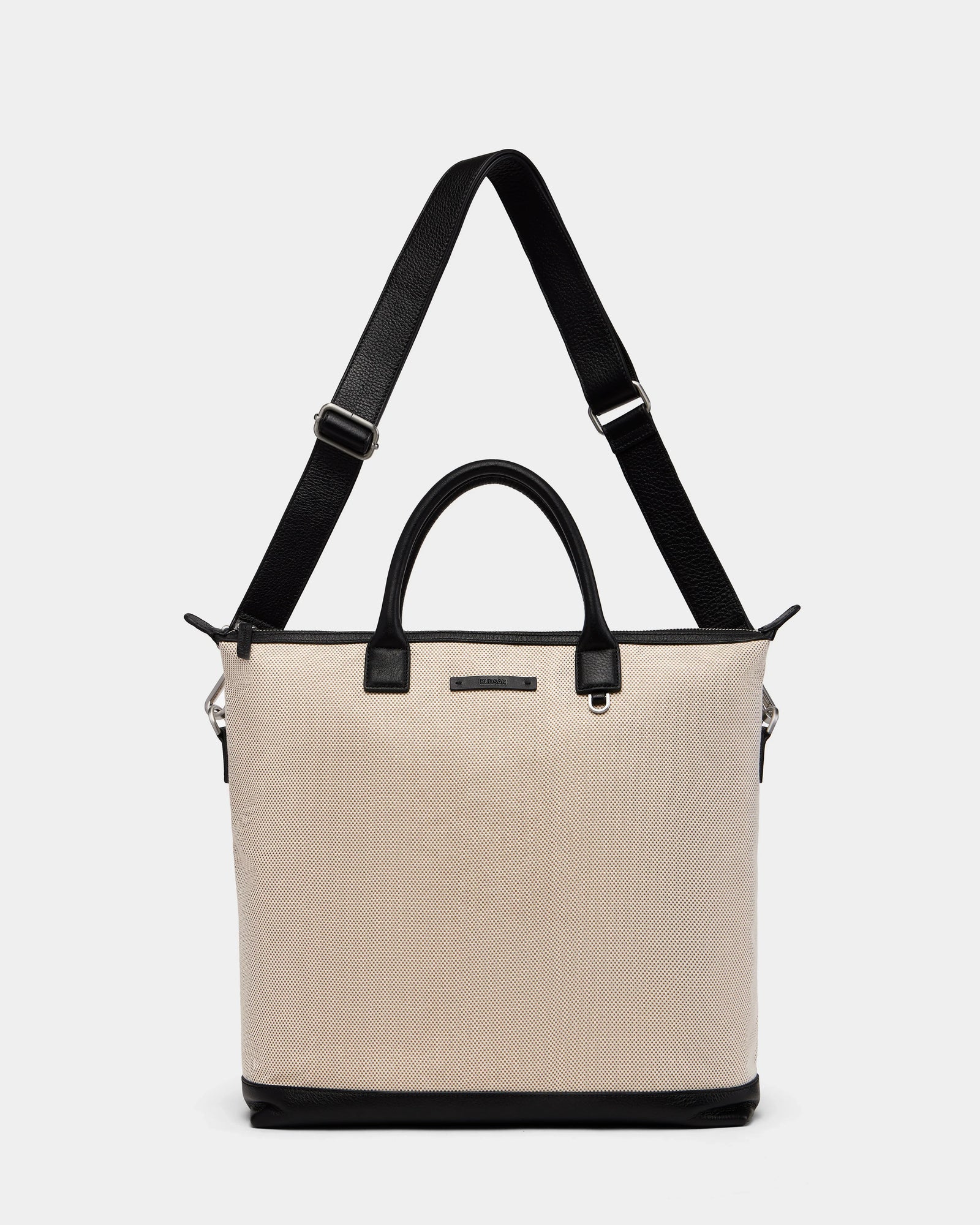 The Rudsak DEXTER - 9324513 Vanilla is a unisex beige canvas tote with black leather handles, a black base, and an adjustable shoulder strap, shown against a plain white background.