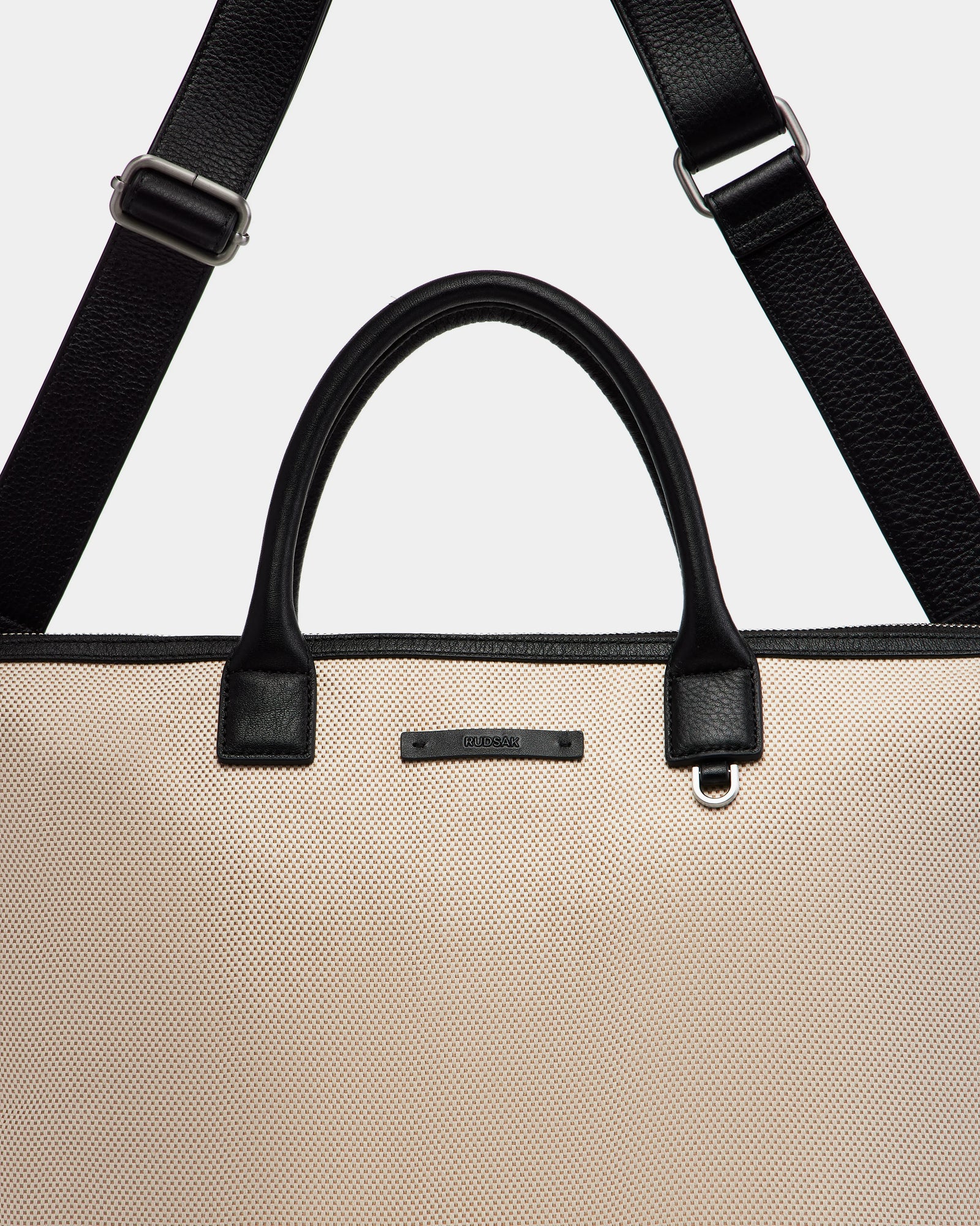 A close-up of the Rudsak DEXTER - 9324513 Vanilla tote bag features beige canvas, black leather handles, and a detachable black shoulder strap, highlighting this versatile accessory against a plain background.