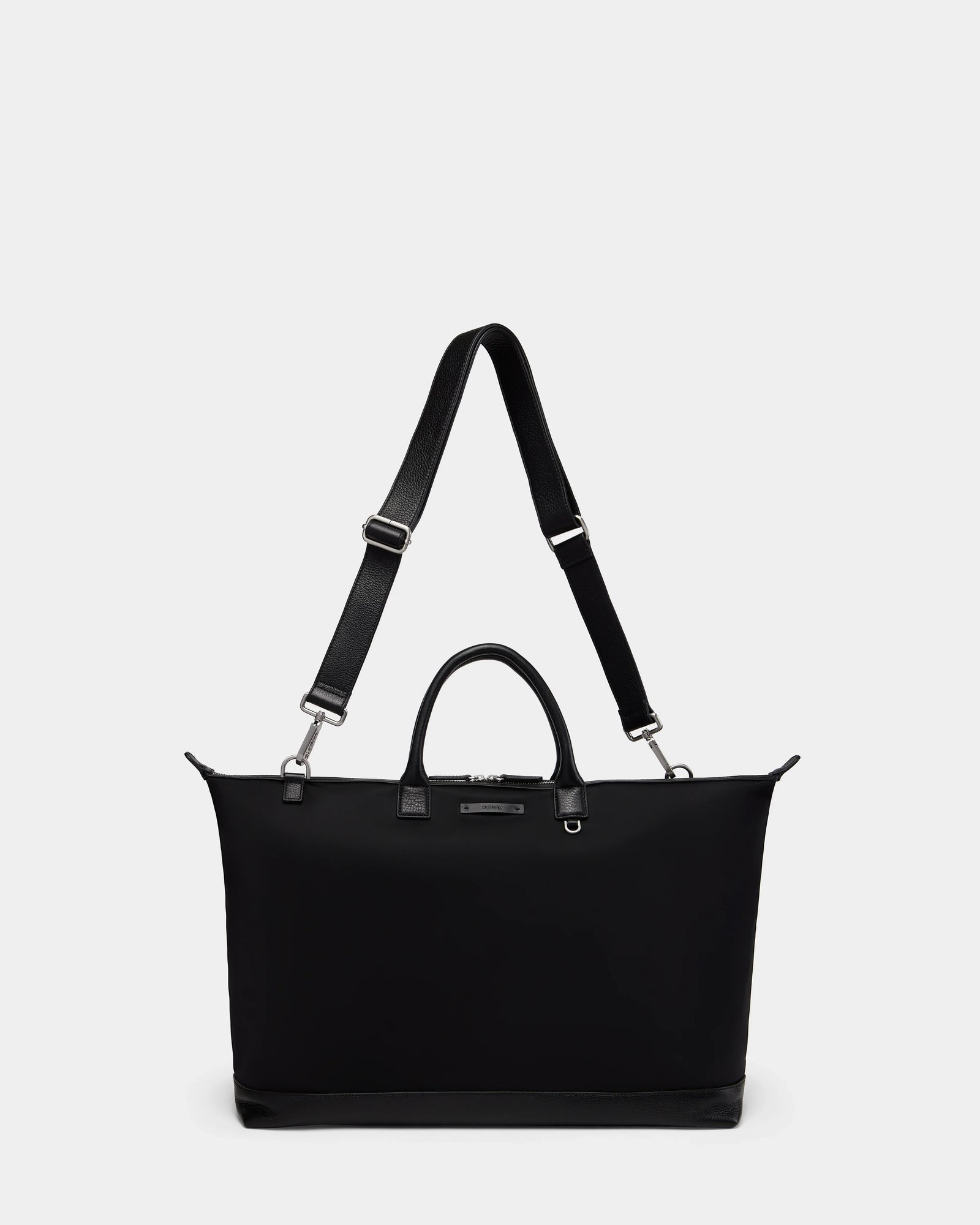The Rudsak DUBLIN - 9324514 Black is a unisex, rectangular canvas leather bag with short handles, a long adjustable strap, silver hardware, and a minimalist design set against a plain white background.