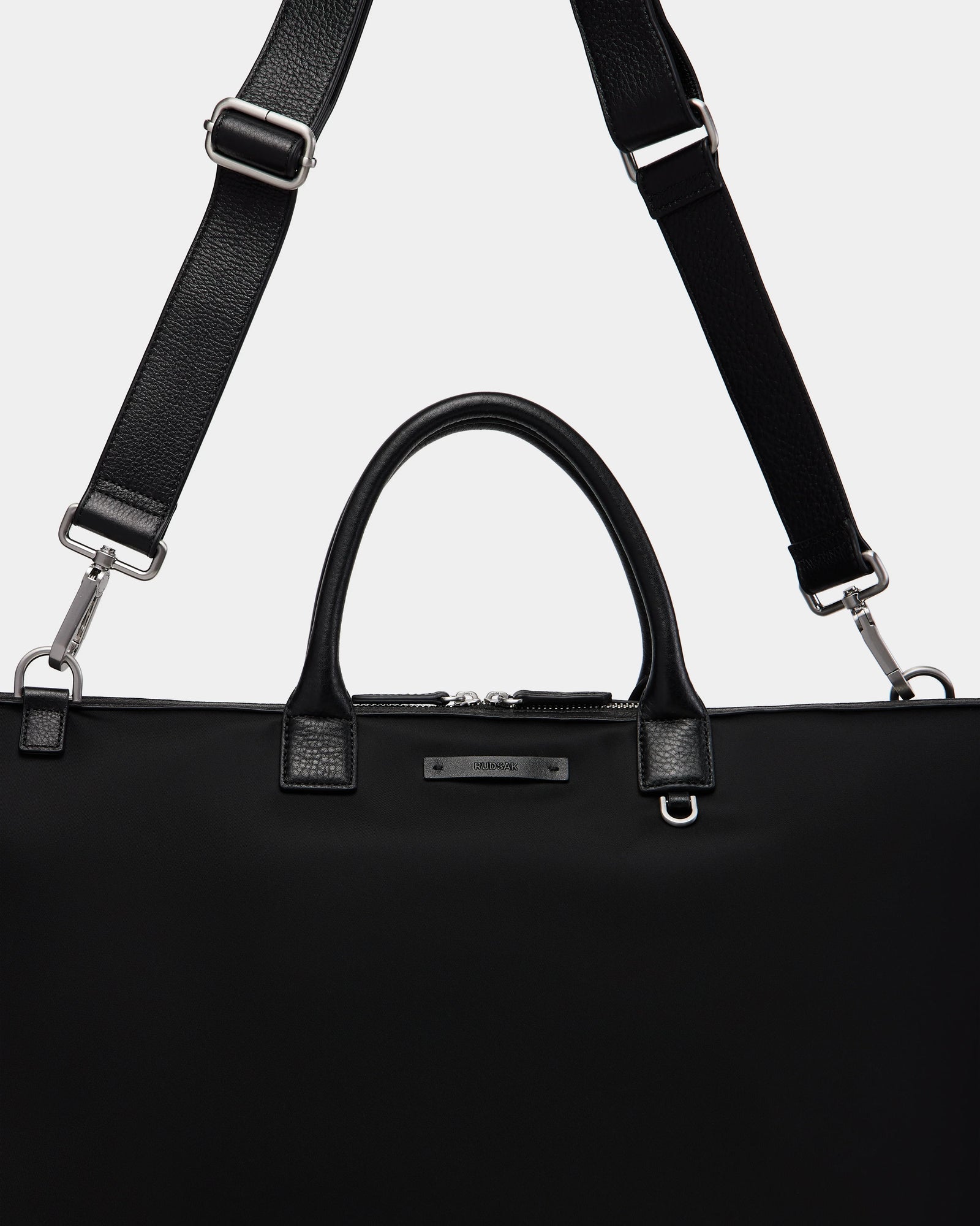 The Rudsak DUBLIN - 9324514 Black is a canvas leather weekender tote with silver hardware, two short handles, and an adjustable, detachable shoulder strap displayed against a plain light background.