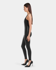 A woman with long brown hair stands in profile against a light gray background, wearing the Rudsak LAWREN - 8523047 Black sleeveless bodysuit and black high-heeled shoes.