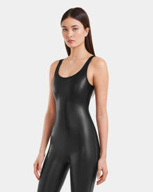 A woman with long straight brown hair wears the Rudsak LAWREN - 8523047 Black fitted, sleeveless vegan leather bodysuit with a shiny finish, standing against a plain white background and looking forward neutrally.