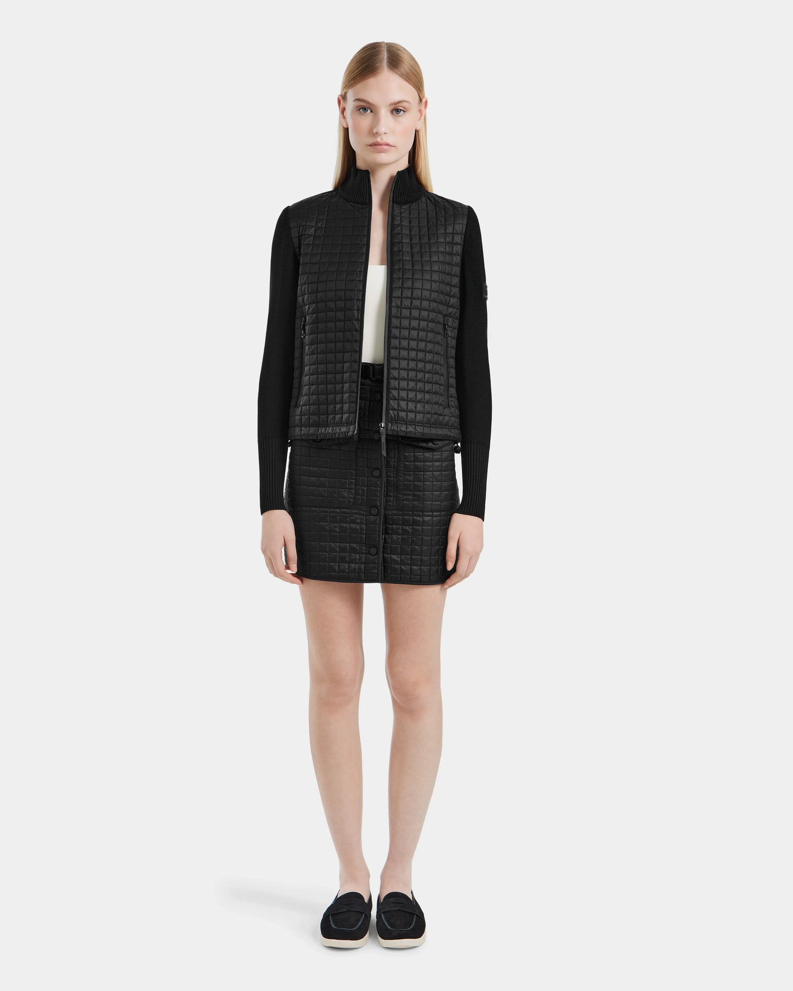 A woman with straight blonde hair and a neutral expression wears the LOUISE - 8124046 Black hybrid knit sweater jacket by Rudsak, styled with a black textured vest, long-sleeve top, skirt with buttons, and black loafers.