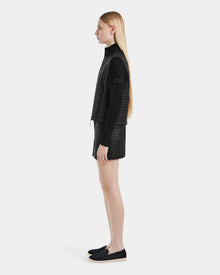 A young woman with long straight blonde hair stands in profile against a white background, wearing the Rudsak LOUISE - 8124046 Black lightweight quilted jacket along with black shorts, a long-sleeve top, and slip-on shoes.