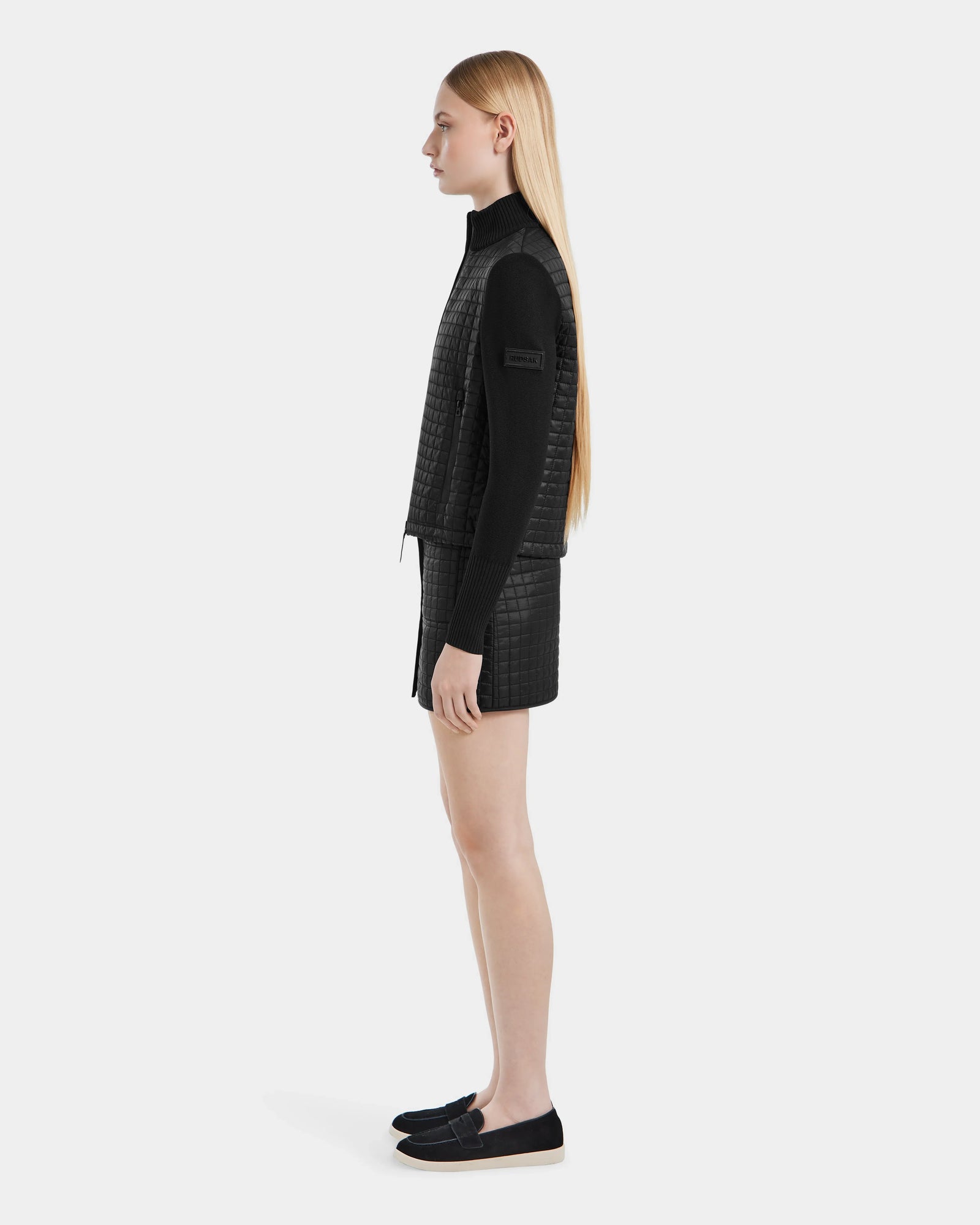 A young woman with long straight blonde hair stands in profile against a white background, wearing the Rudsak LOUISE - 8124046 Black lightweight quilted jacket along with black shorts, a long-sleeve top, and slip-on shoes.
