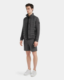 A man stands against a white background wearing the Rudsak RAGAB - 6124577 Grey hybrid jacket with lightweight down insulation, matching zip-up, grey shorts, and white sneakers—perfect for transitional weather. He looks slightly to the side, hands relaxed.