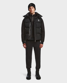 A person faces forward against a white background, wearing Rudsak’s REN M - 9123512 Black puffer vest over a black zip-up jacket, paired with black jogger pants, boots, and a black beanie.
