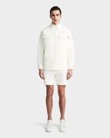 A man faces forward wearing the Rudsak KERRY - 9124003 Eggshell M quilted shirt jacket with eco-friendly insulation, paired with white shorts and sneakers, against a plain light gray background.