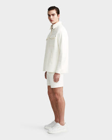 A young man stands sideways wearing the Rudsak KERRY - 9124003 Eggshell M unisex quilted shirt jacket with eco-friendly insulation, matching shorts, and white sneakers. He faces left and looks at the camera against a plain light gray background.