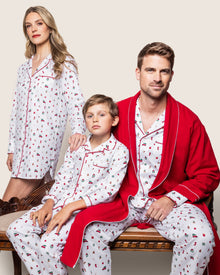 A family of three sits on a bench in matching Petite Plume Kids Twill Pajama Sets in Winter Nostalgia, adorned with festive prints and red trim. The man adds a red robe. A neutral backdrop enhances the cozy scene, poised for the gentle chime of Christmas bells to complete their cheerful image.