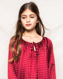 A young girl with long brown hair wears Petite Plumes Girls Silk Delphine Nightgown in Bordeaux Polka Dot. She stands against a light, textured background, looking slightly to the side with a calm expression.