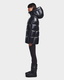 A person in profile wears the Rudsak RITA S - L123527 Black womens down puffer jacket with a large hood, paired with black leggings and black winter boots, set against a plain light background.