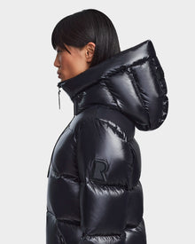 A person with straight black hair, seen in profile, wears the Rudsak RITA S - L123527 Black jacket with a large hood, RDS-certified down, and an R arm patch. The shiny weather-resistant coat stands out against a plain white background.