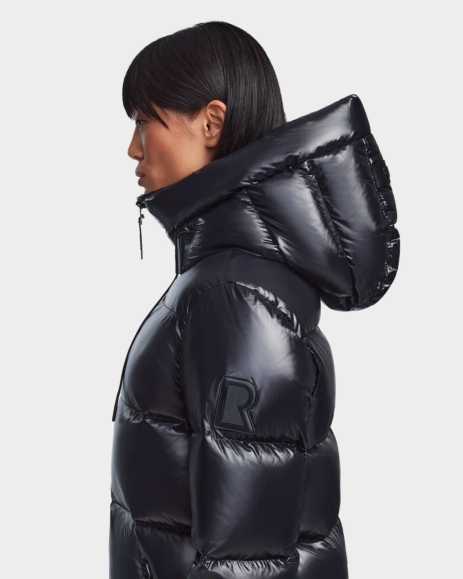 A person with straight black hair, seen in profile, wears the Rudsak RITA S - L123527 Black jacket with a large hood, RDS-certified down, and an R arm patch. The shiny weather-resistant coat stands out against a plain white background.