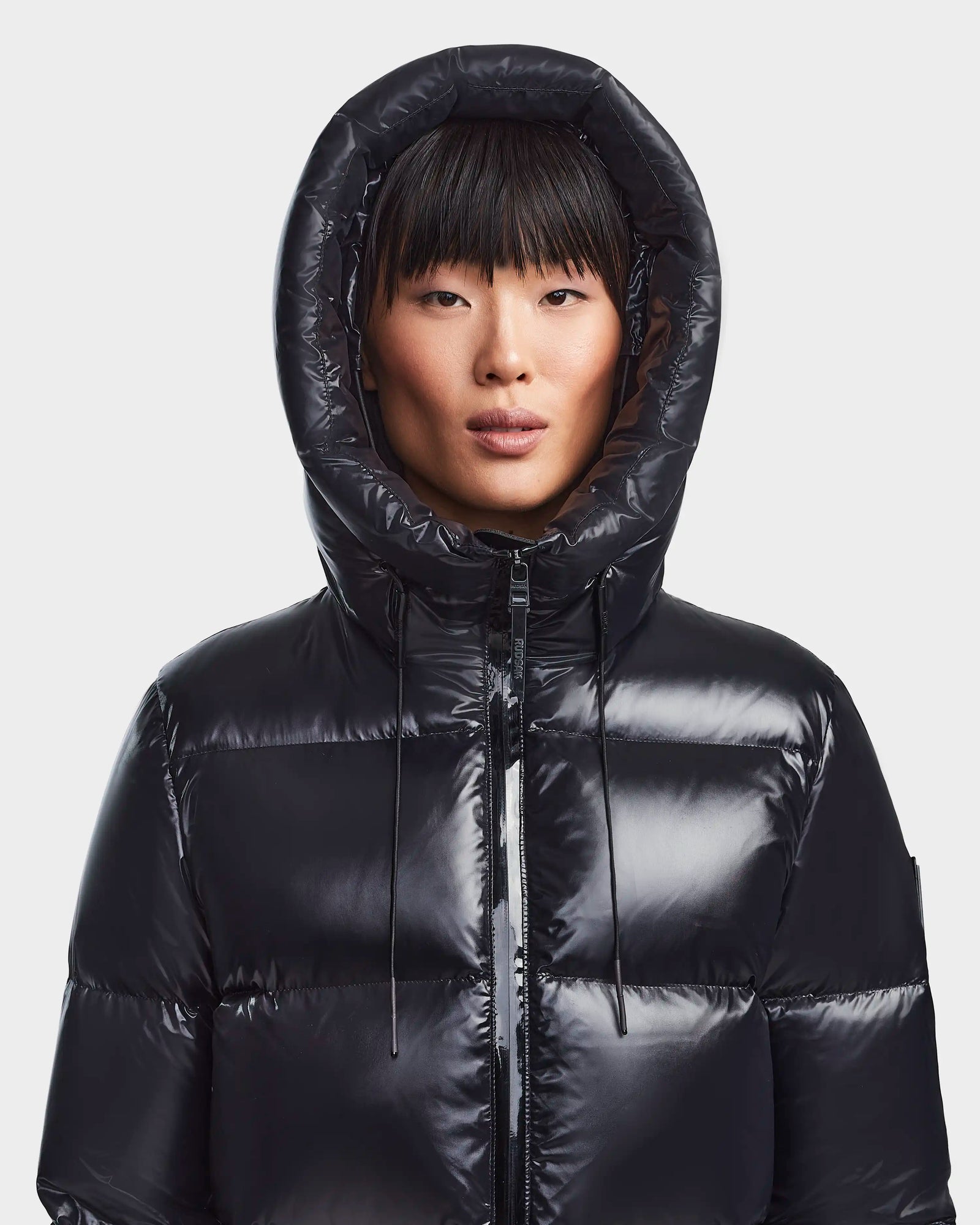 A person in the Rudsak RITA S - L123527 Black weather-resistant puffer with the hood up looks directly at the camera against a plain light background.