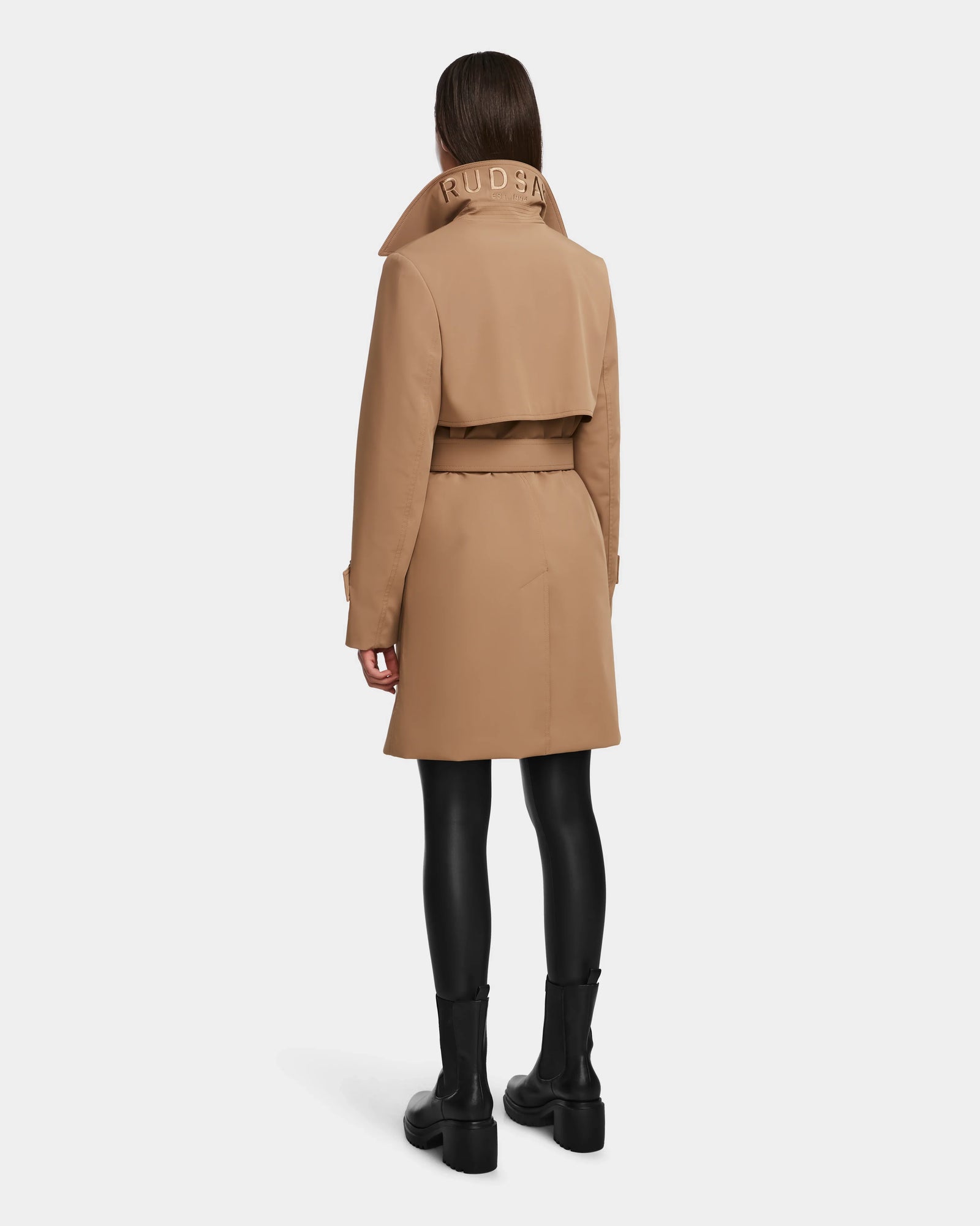 A person faces away in the Rudsak KIMBERLY - 8124801 Camel trench, featuring a tan water-repellent finish, belted waist, vented back, and RUDSAK on the collar. Styled with black leggings and ankle boots against a plain white background.