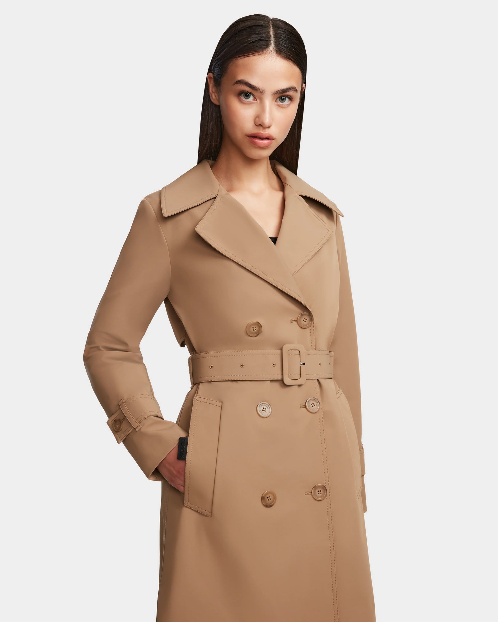 A woman with straight, dark hair models the Rudsak KIMBERLY - 8124801 Camel trench coat with a belted waist and pockets, standing against a plain white background.