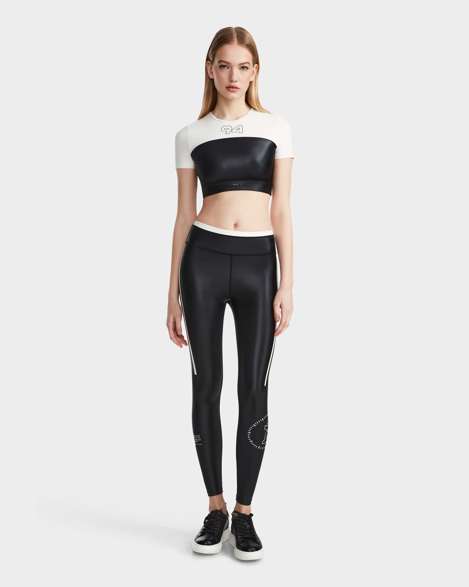 A woman with straight light brown hair stands facing forward, wearing Rudsak’s LILIANE - 8524018 Black women’s fitted crop top and matching high-waisted black leggings with white stripes, paired with black sneakers, against a plain light gray background.