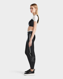 A woman with long blonde hair stands sideways, wearing the Rudsak TAYLA - 8524015 Black short-sleeve crop top, high-performance leggings with a white logo on the calf, and black platform sneakers against a plain light background.