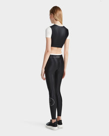 A woman with long, straight hair wears the Rudsak TAYLA - 8524015 Black crop top and matching high-waisted leggings featuring a small white circular logo, paired with black platform sneakers, standing against a plain background.
