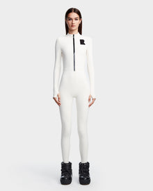 A woman models the Rudsak DAVINA - 8523534 Iceland: a sleek, white, long-sleeve jumpsuit with front zipper and black R on the chest, styled with chunky black boots against a plain white background for an après-ski look.