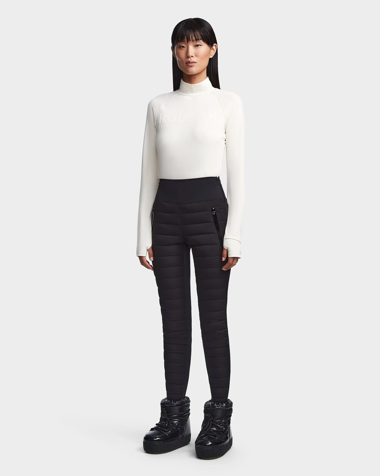 A woman with straight black hair and a neutral expression wears the Rudsak KILTON - 8523533 Iceland fleece-lined jersey top and black quilted high-waisted pants, paired with chunky boots, against a plain light gray background. Ideal for winter.