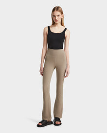 A woman with straight blonde hair and a neutral expression wears the Rudsak ADRIANA - 8524009 Cement high-waisted taupe flared-leg leggings, a black tank top, and black slide sandals against a plain white background.