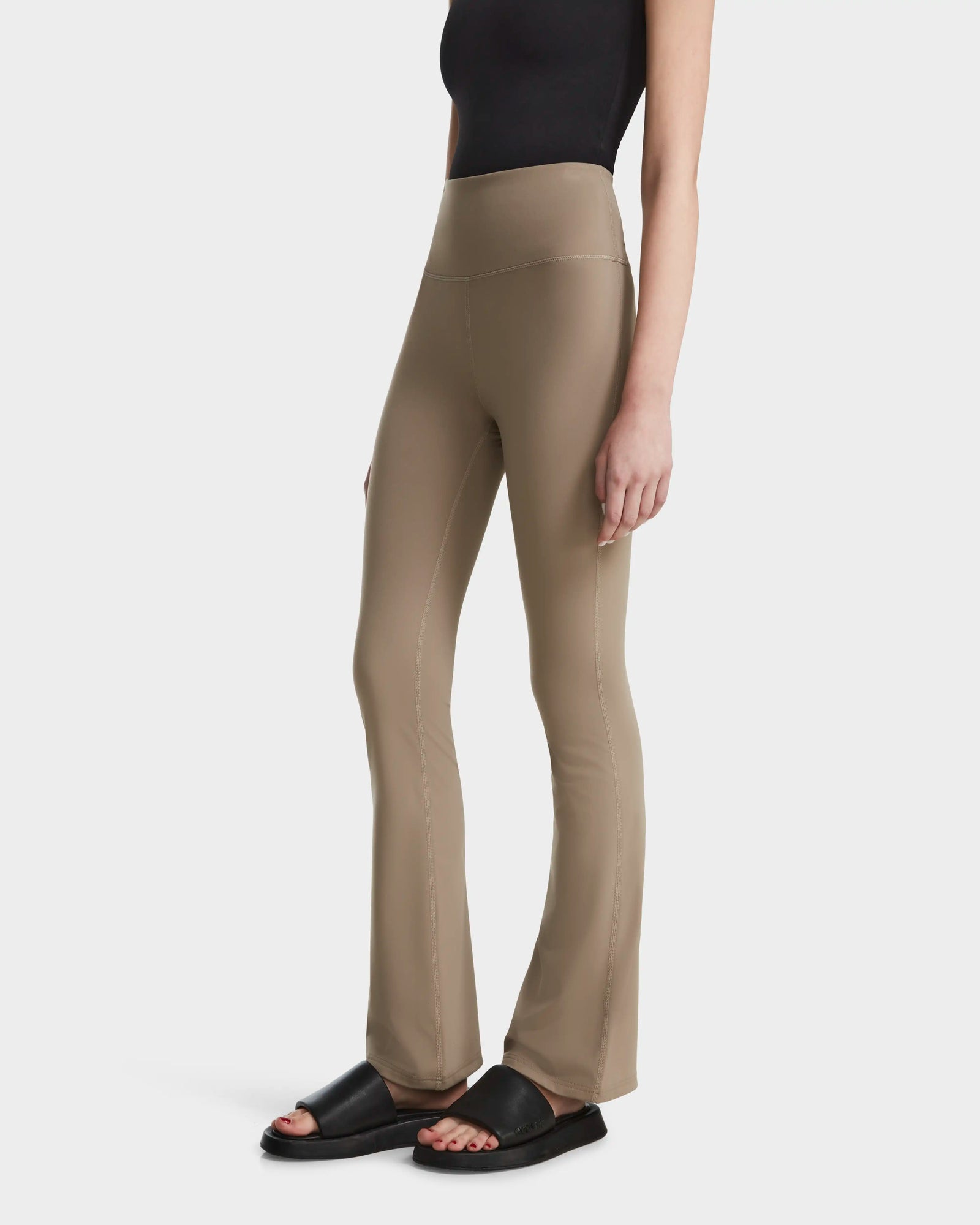 A person wearing the Rudsak ADRIANA - 8524009 Cement taupe high-waisted flared-leg leggings, a black sleeveless top, and black slide sandals stands against a plain white background.